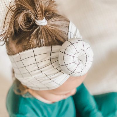A little girl wearing a Charlie - Bamboo Baby Knotted Headwrap by Tiny Knot Co with a checkered pattern.
