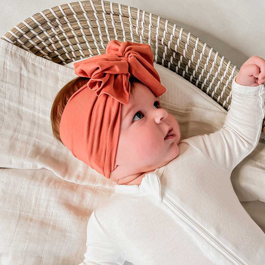 A baby wearing a headband is peacefully resting on a wicker bed from Tiny Knot Co.